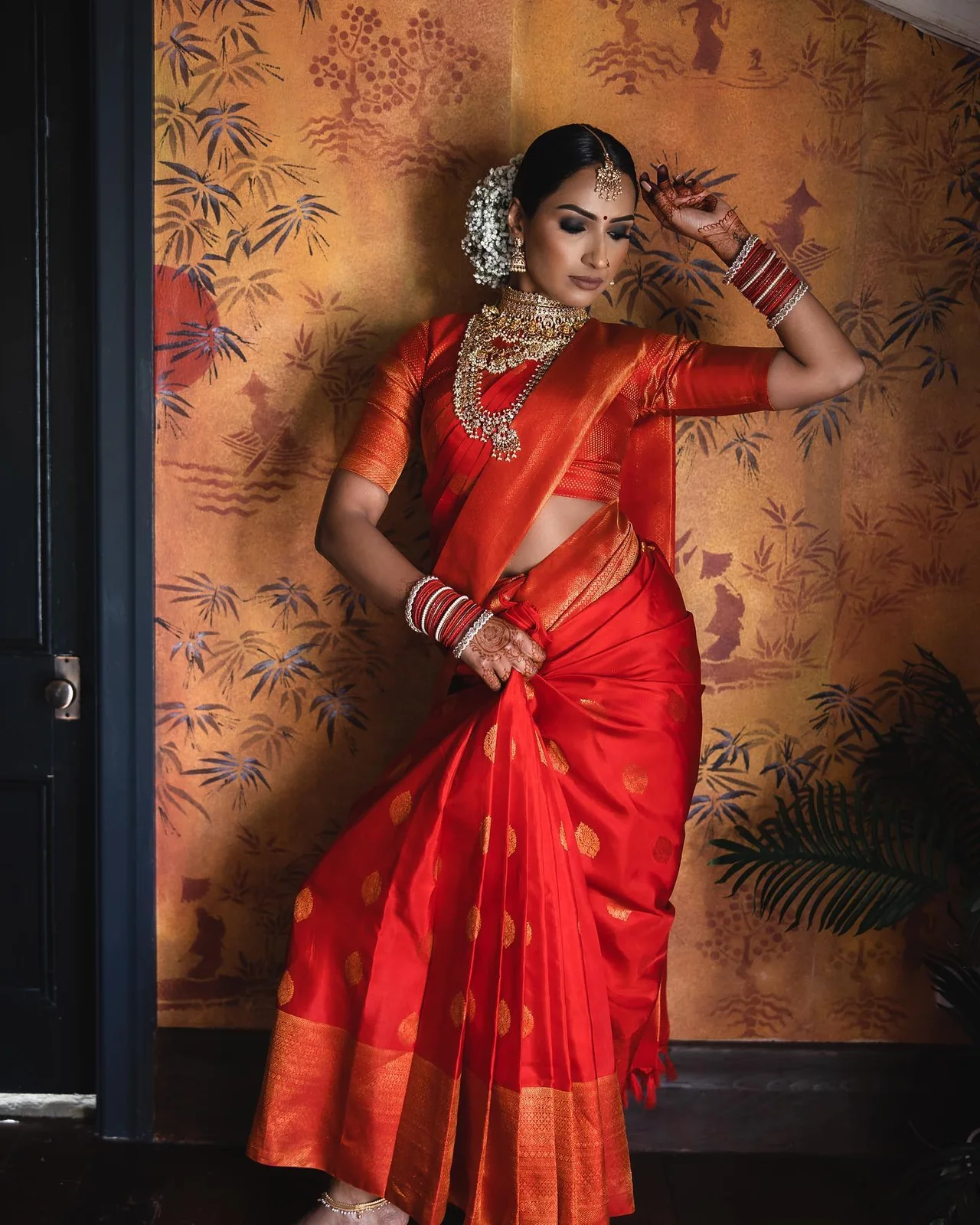 Full-length editorial of a bride in a red Kanjeevaram silk saree against a hand-painted chinoiserie wall.