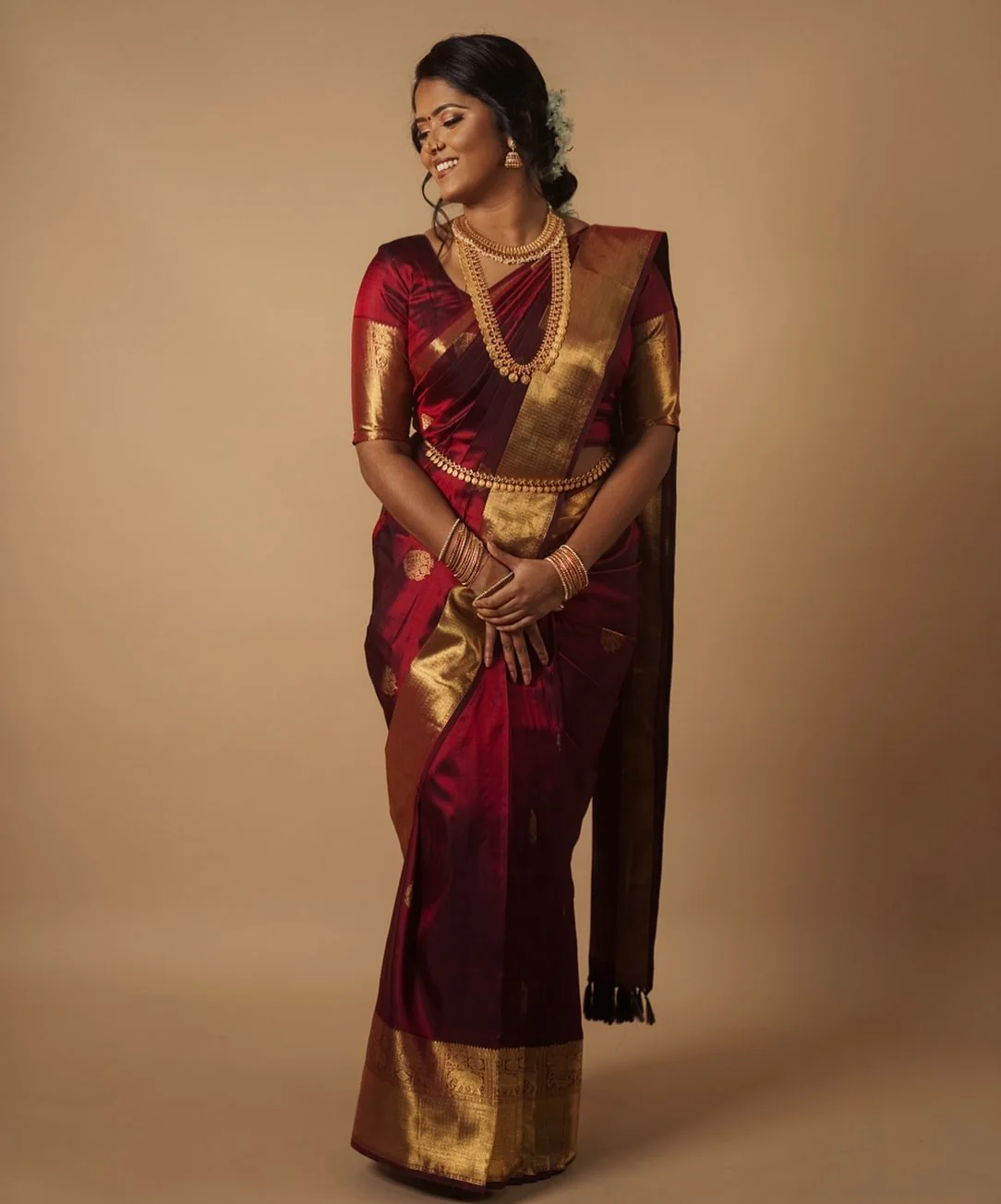 Studio portrait of a woman in a maroon and gold Kanjeevaram silk saree with a layered gold haram, on a warm ground.