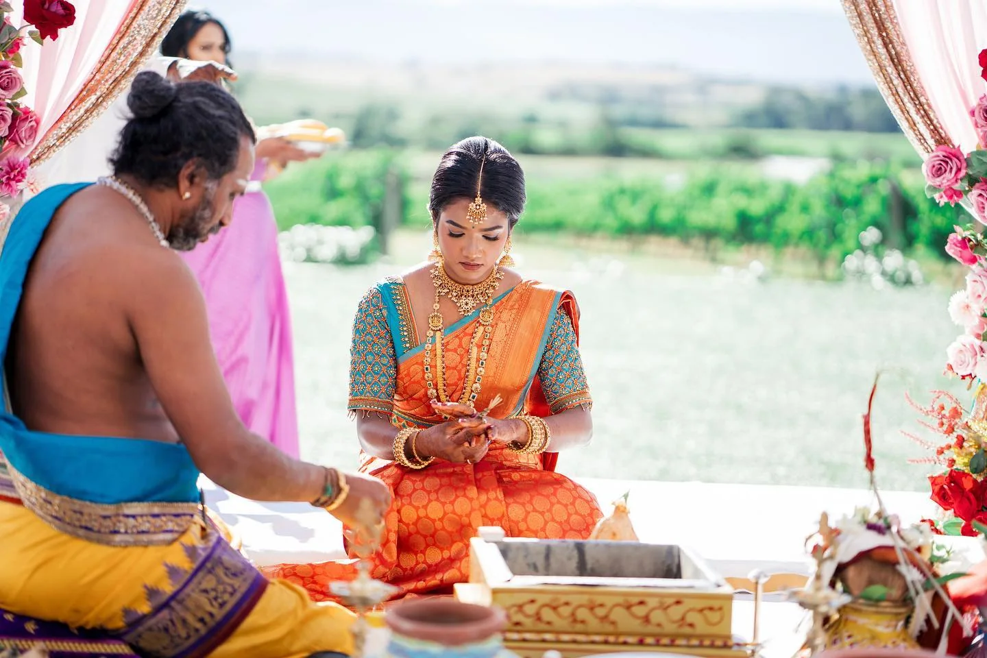 Bride in an orange Kanjeevaram silk saree at an outdoor wedding function, hands together in a ritual moment.