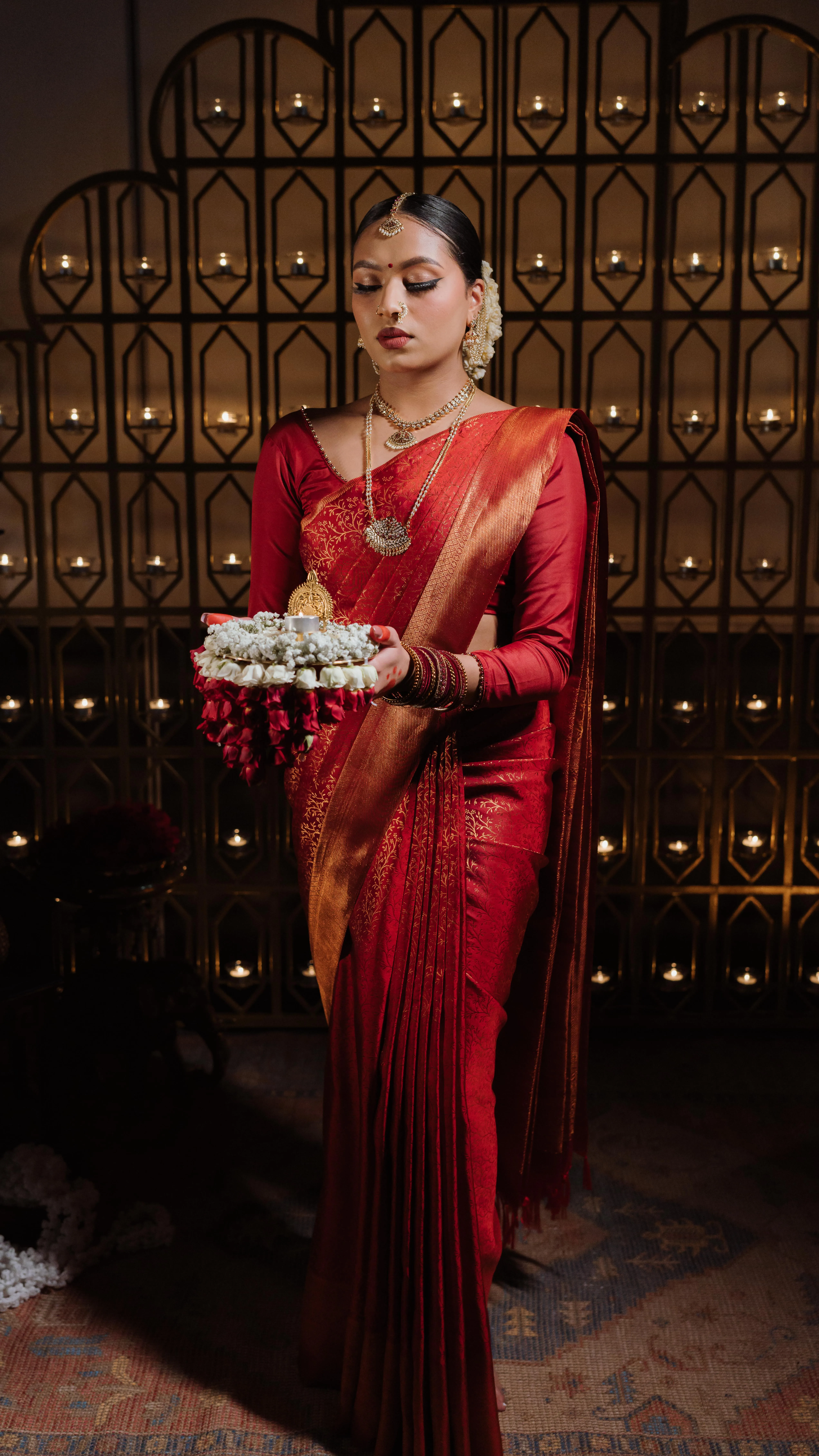 Bride in temple-red Kanjeevaram holding a brass lamp before an arched wall of tea-lights, jasmine and marigold.