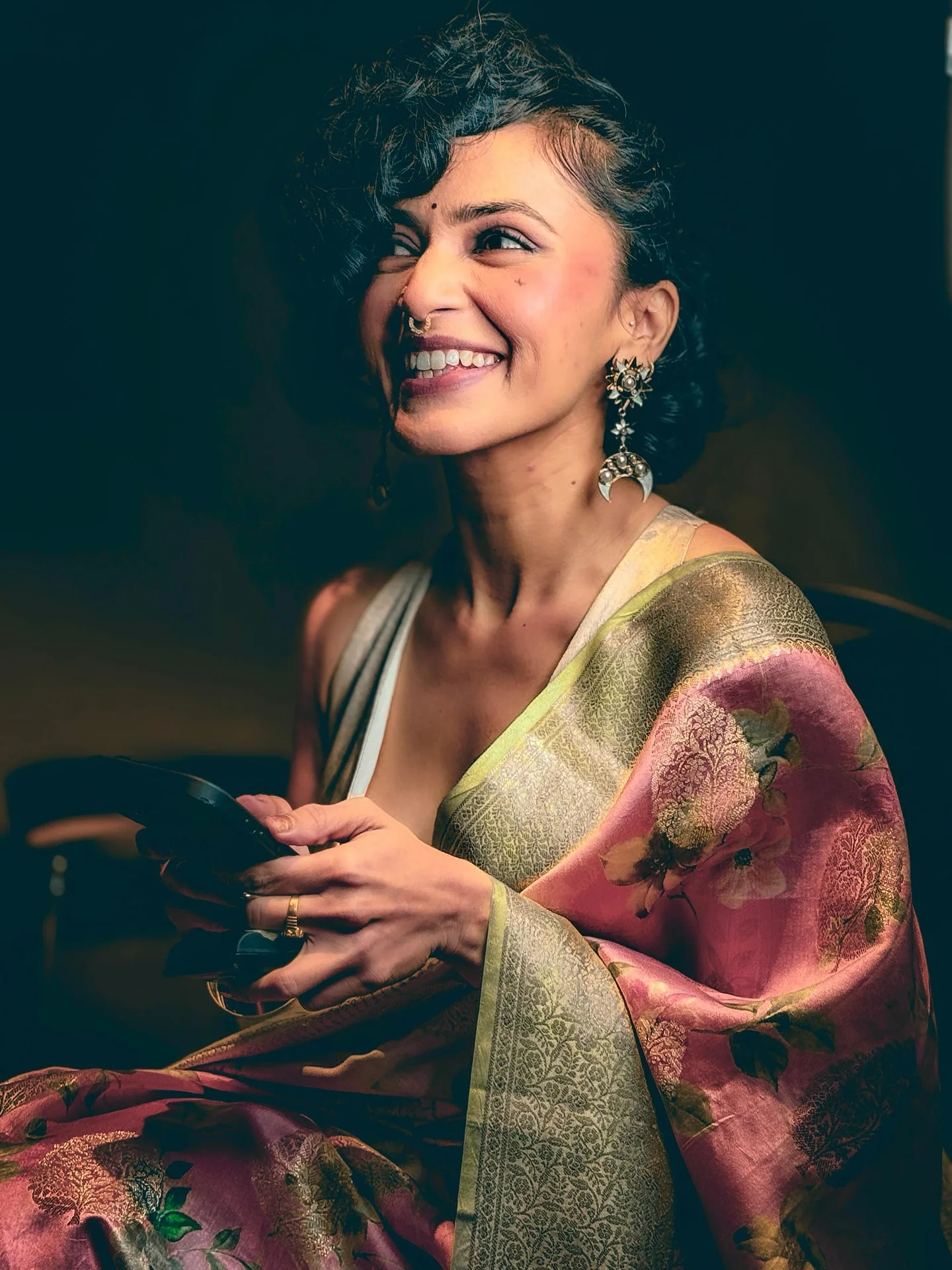 A woman in pink-gold floral silk saree with gold jhumkas, laughing against a warm dark ground.