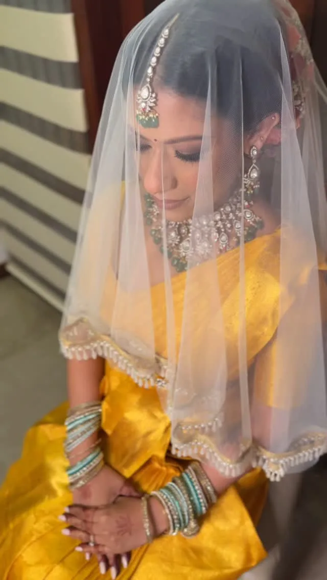 Bride in yellow silk saree with white veil, diamond and emerald bridal jewellery, green bangles and mehndi.