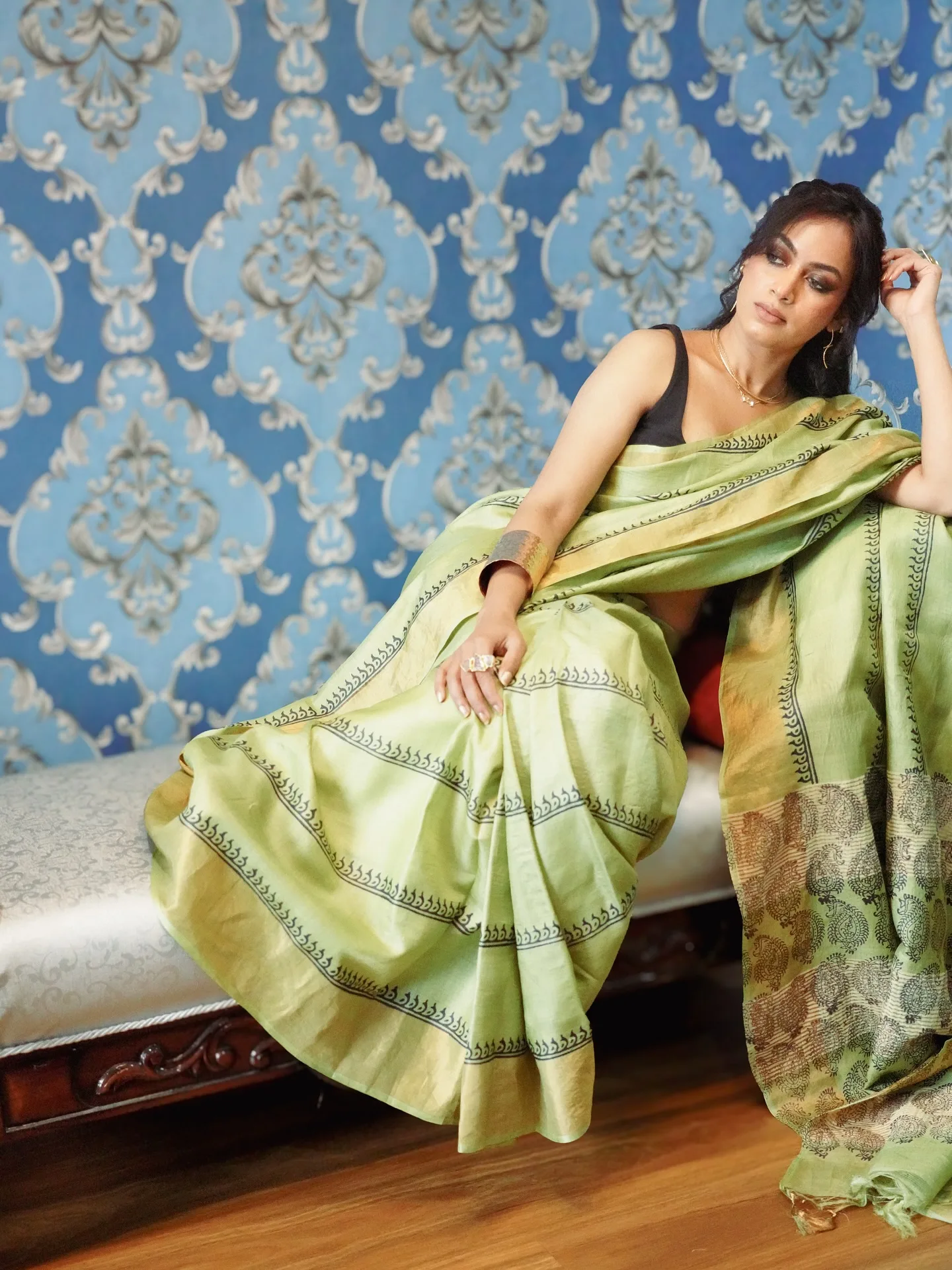 Woman reclining in a green tussar-silk saree against an ornate blue damask wall.