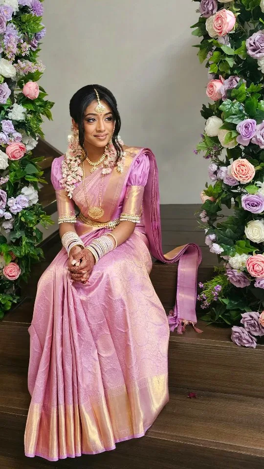 Bride in pink-lilac Kanjeevaram between pastel floral pillars with jasmine and mehndi.