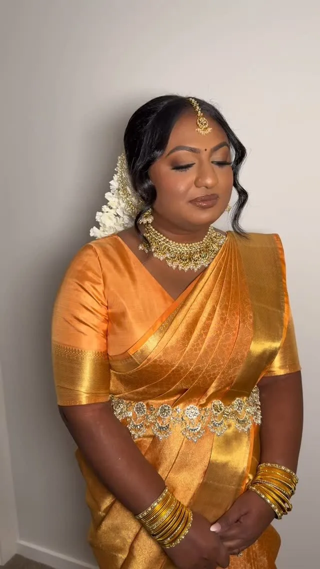 Bride in orange-gold silk saree with jasmine gajra and gold jewellery.