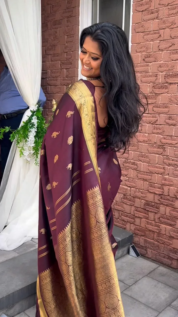 Woman from behind, showing a maroon silk saree back with gold elephant motifs, outdoor setting with a jasmine garland.