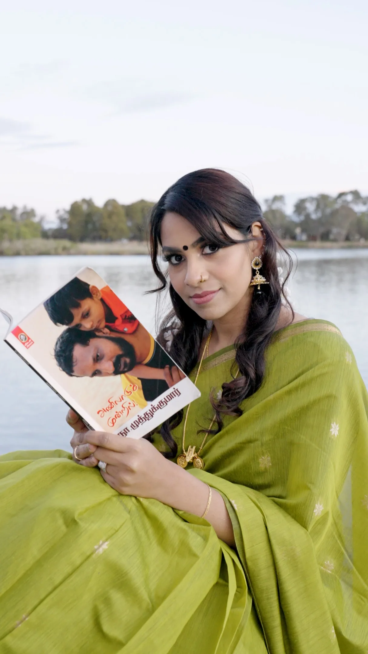 Woman in green cotton-silk saree styled by a suburban Melbourne lake, holding a Tamil-print book.