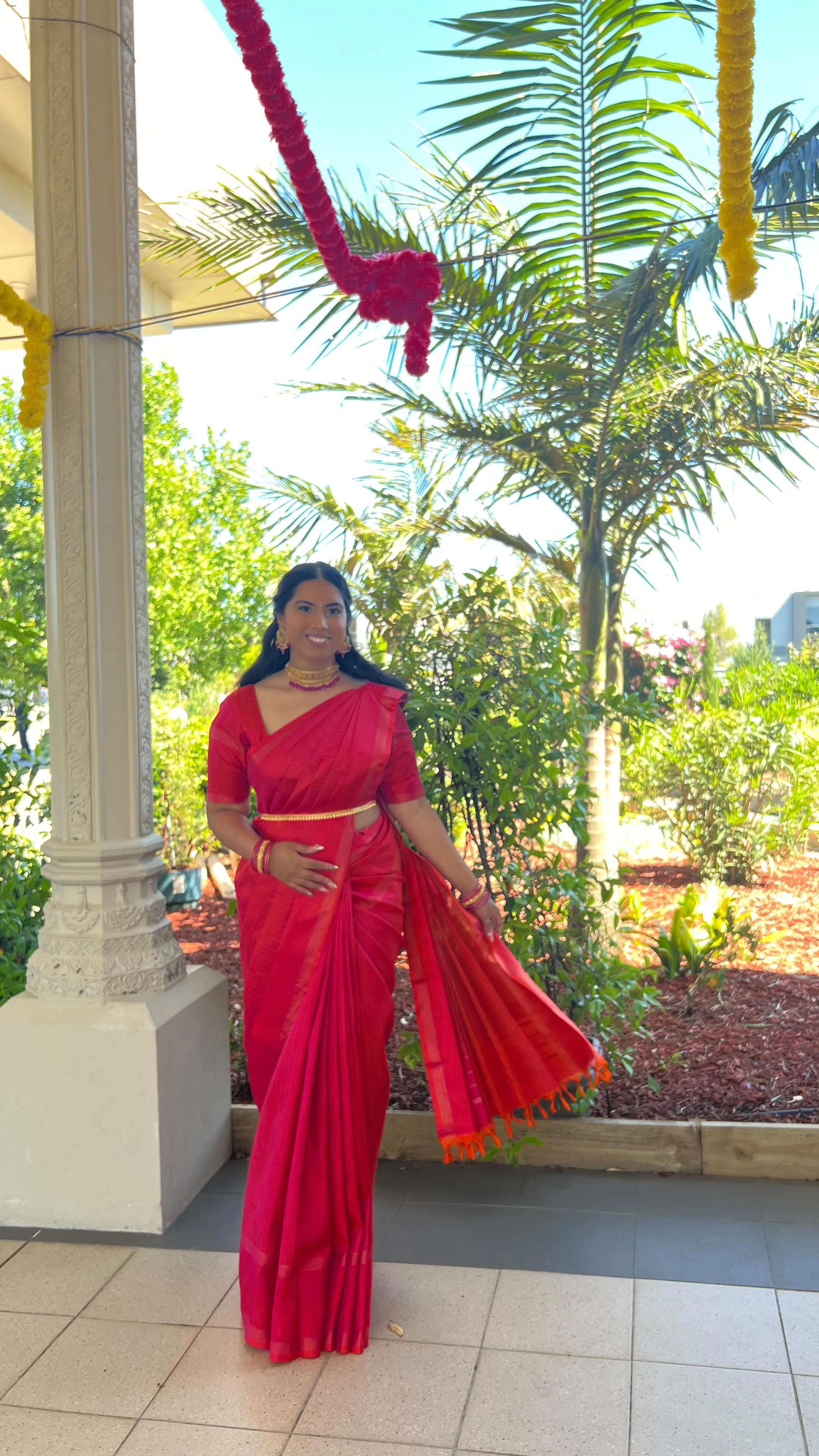 Woman in red silk on a columned garden veranda with marigold garlands, palm and landscaping behind her.