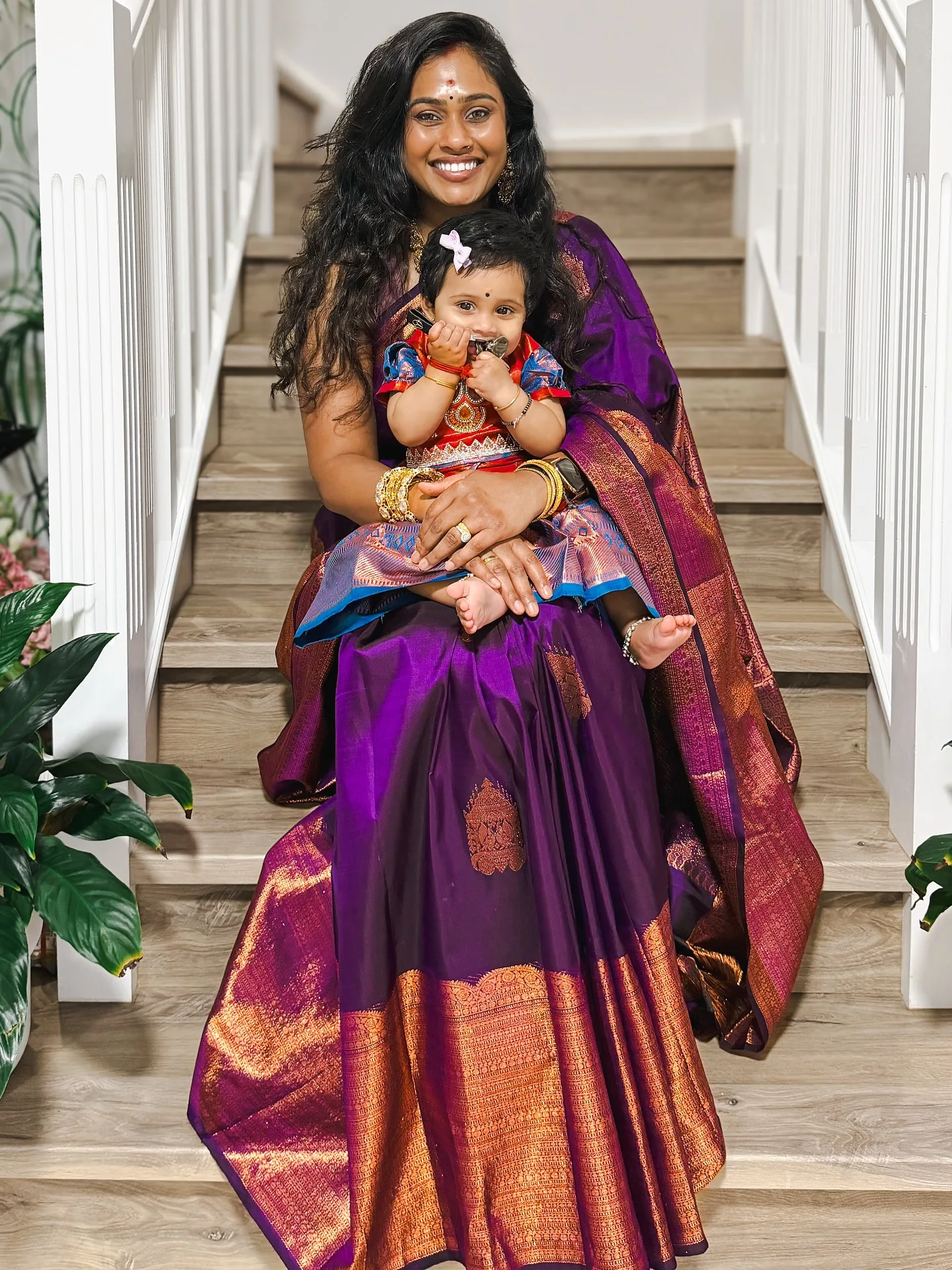 Mother in purple Kanjeevaram on stairs holding a baby dressed in a matching pattu pavadai — a family function.