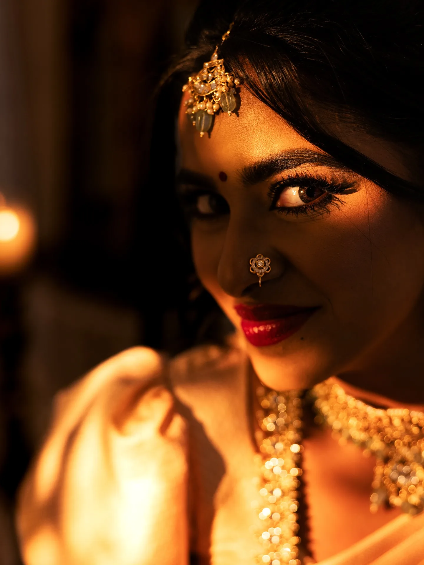 Moody bridal beauty close-up — maang tikka, nose ring, red lips, warm candlelight.