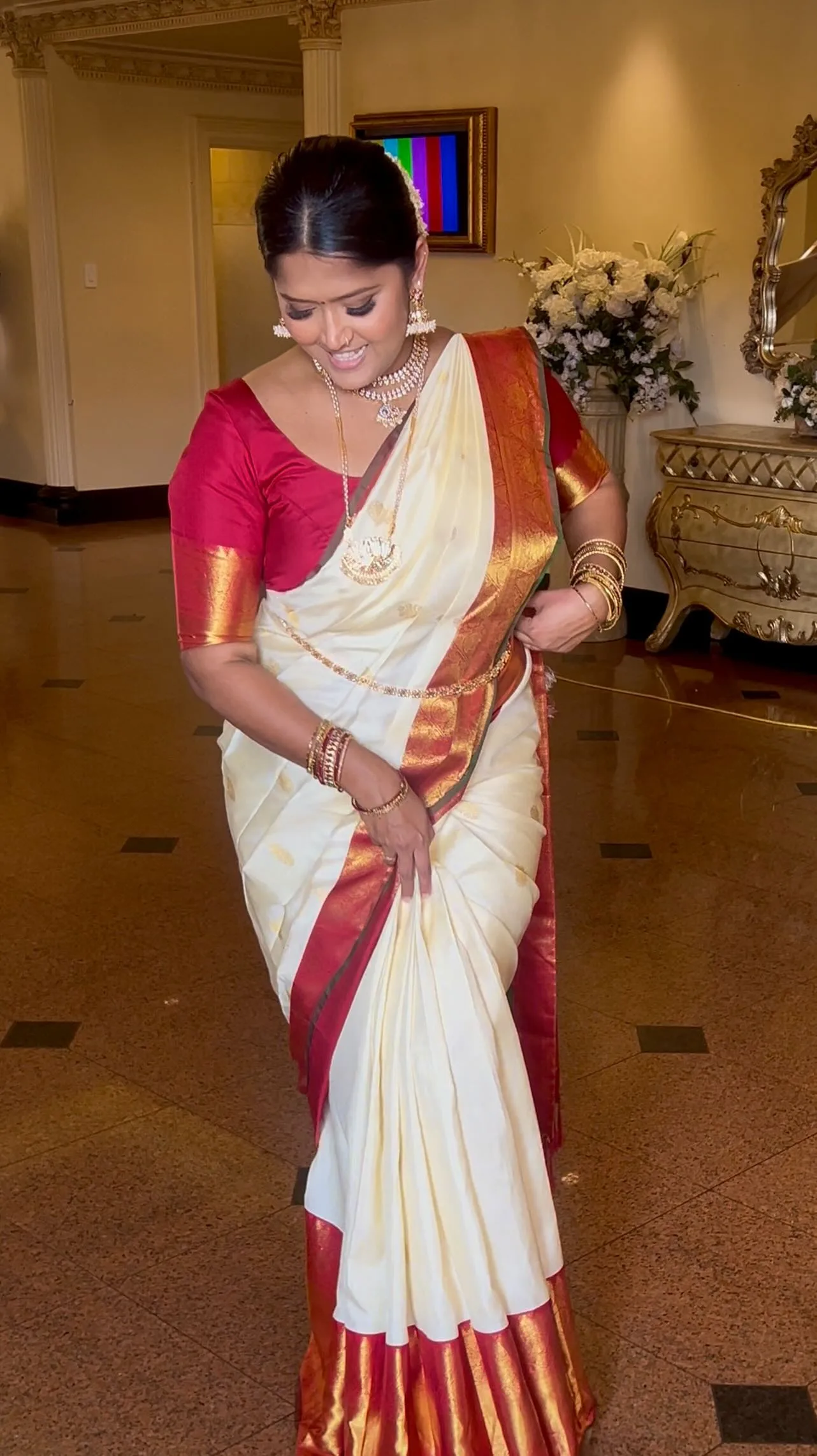 Woman in cream kasavu silk with red-gold border, adjusting her drape in a gilded hotel lobby.